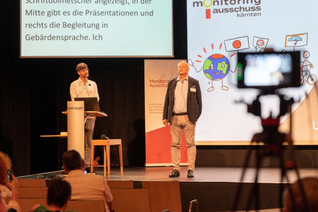 Heinz Pfeifer und Tobias Buchner stehen auf der Bühne. Im Hintergrund sieht man zwei Bildschirme. Links den Schriftdolmetsch und rechts einen Teil der Powerpointpräsentation. Rechts vorne im Bild sieht man eine Kamera, die auf die beiden gerichtet ist.