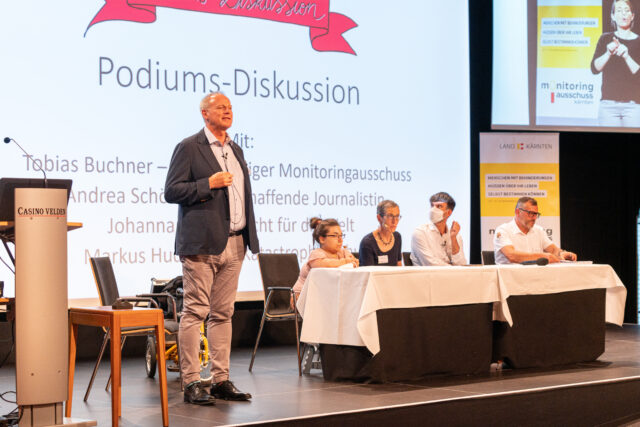Heinz Pfeifer kündigt die Podiums-Diskussion an. ER steht links neben dem Podium. Andrea Schöne, Johanna Mang, Tobias Buchner und Markus Hudobnik diskutieren mit dem Publikum. Hinter dem Podium sieht man die Powerpoint-Folie auf der Podiums-Diskussion steht.