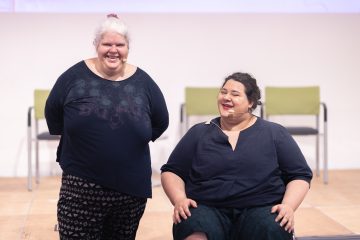 Moderatorinnen Iris Kopera und Yuria Knoll lachen in die Kamera. Beide tragen dunkle Kleidung und nutzen ein Headset als Mikrofon.