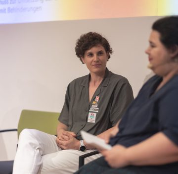 Nahes Portrait von Katrin Neudolt am Podium, verschwommen sieht man Moderatorin Yuria Knoll