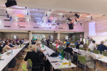 Von der Seite sieht man den gefüllten Saal des ÖGB und auch das Podium, Tobias Buchner und Daniela Rammel halten ihren Startvortrag.