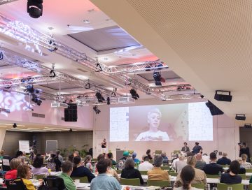 Blick von hinten über die Teilnehmenden hinweg auf die Bühne, man Sieht Laura Botschaft am großen Bildschirm. am Podium sitzen Katrin Neudolt und Moderatorin Yuria Knoll.