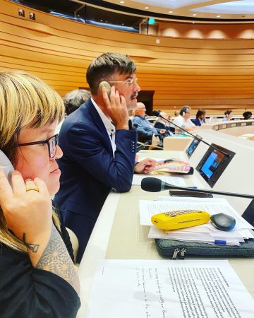 Rammel und Buchner halten Mikrofone an ihr Ohr, sie sitzen im Sitzungssaal des UN-Fachausschuss auf ihren Arbeitsplätzen