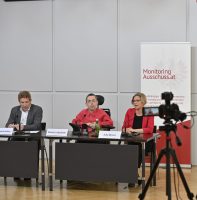 3 Personen auf Rednerpult bei Pressekonferenz, Kamera filmt sie