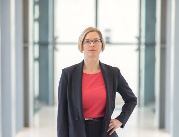 Frau mit kinnlangen blonden Haaren und Brille in Blazer mit Hand in der Hüfte