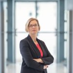 Frau mit kinnlangen blonden Haaren und Brille in Blazer verschränkt Arme