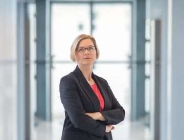 Frau mit kinnlangen blonden Haaren und Brille in Blazer verschränkt Arme