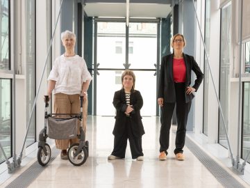 3 Frauen in hellem Gang: von links ältere Frau mit Rollator,kleinwüchsige Frau mit Vokuhila, Frau mit kurzen blonden Haaren und Blazer