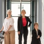 3 Frauen in hellem Gang: von links ältere Frau mit Rollator, Frau mit kurzen blonden Haaren und Blazer, kleinwüchsige Frau mit Vokuhila