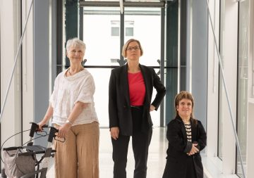 3 Frauen in hellem Gang: von links ältere Frau mit Rollator, Frau mit kurzen blonden Haaren und Blazer, kleinwüchsige Frau mit Vokuhila