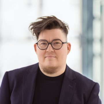 Person mit kurzen Haaren, Brille, Bart und dunklem Blazer