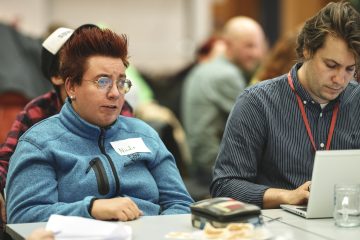 Rollifahrerin mit roten Haaren und Brille diskutiert an Tisch, rechts tippt ein junger Mann am Laptop