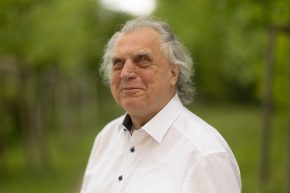 Erich Schmid blickt seitlich in die Ferne und lächelt leicht. Er hat weiß-graue schulterlange Haare und trägt ein weißes Hemd. Am Bild sind Kopf und oberer Oberkörper. Im Hintergrund sind verschwommene grüne Bäume.