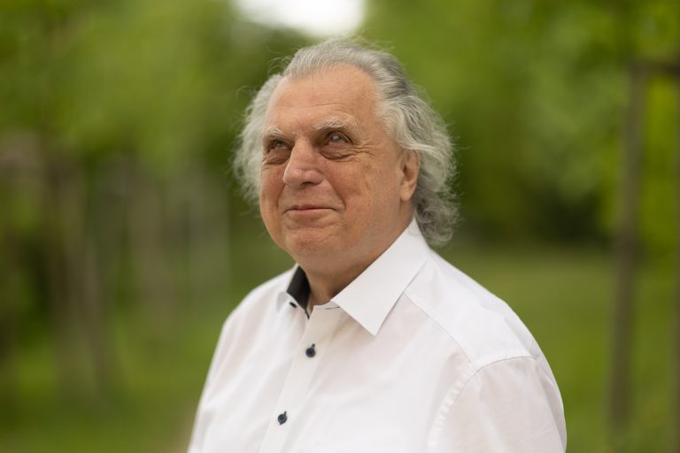 Erich Schmid blickt seitlich in die Ferne und lächelt leicht. Er hat weiß-graue schulterlange Haare und trägt ein weißes Hemd. Am Bild sind Kopf und oberer Oberkörper. Im Hintergrund sind verschwommene grüne Bäume.